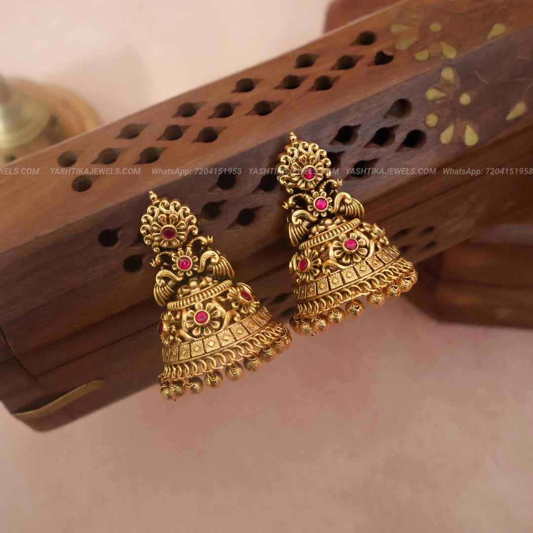 A pair of traditional Indian gold-toned jhumka earrings with small pink gemstone accents, displayed on a carved wooden block against a soft beige background.