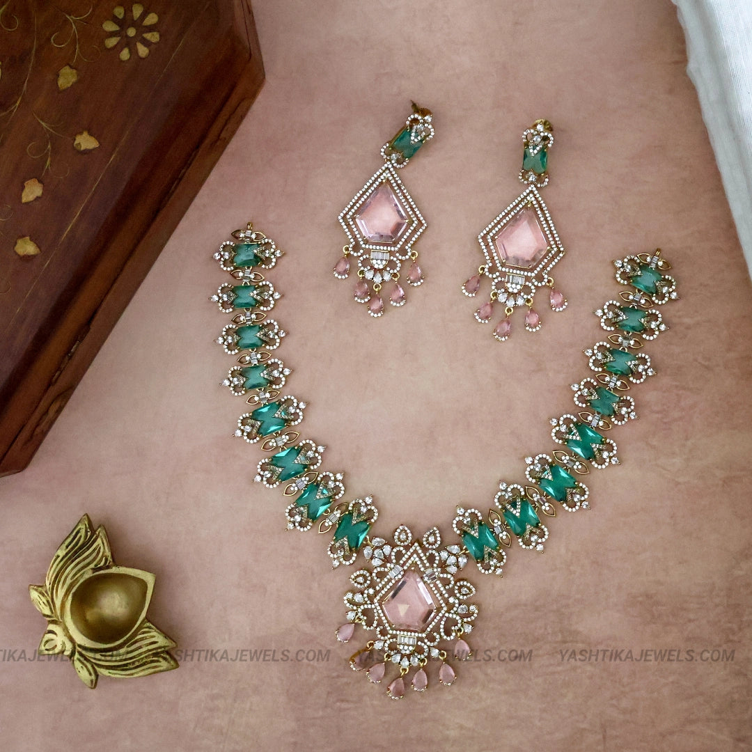 Gold-plated jewelry set featuring a necklace and matching earrings with pink stones and green stones, displayed on a skin-toned surface next to a wooden box.