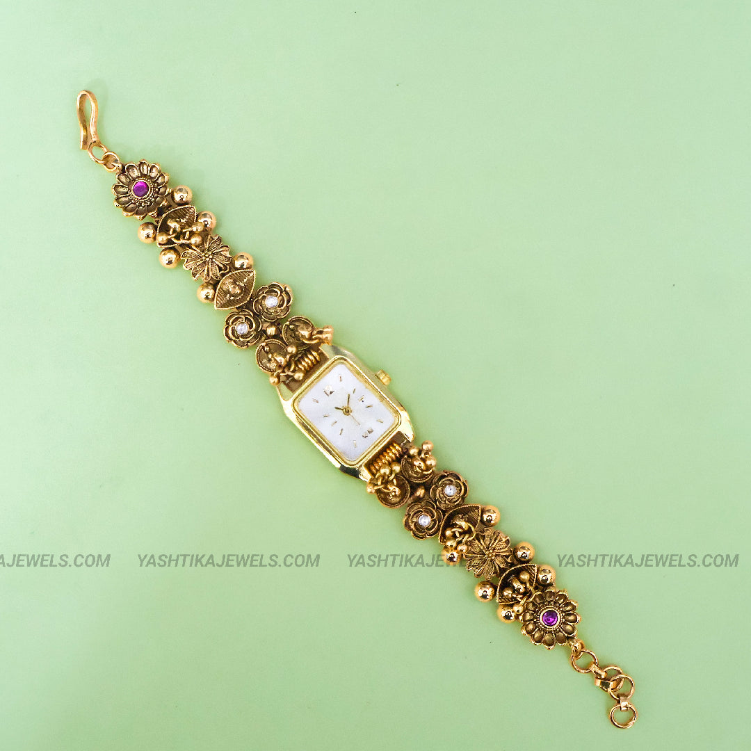 An ornate gold-plated bracelet watch with a square white dial, decorated with floral motifs, small beads, and purple gemstone accents, displayed diagonally on a light green background.