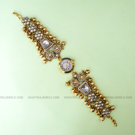 An ornate gold-plated traditional Indian Kundan wrist watch bracelet with a round watch face at the center, embellished with clear stones and gold beads, shown on a light green background.