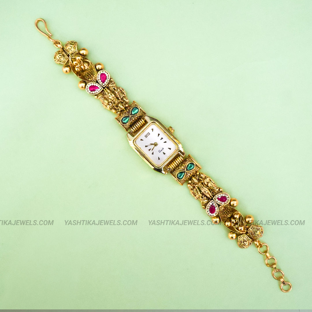 An ornate gold-plated watch bracelet with a rectangular white dial, decorated with pink gemstone accents and small green stone details, shown on a light green background.