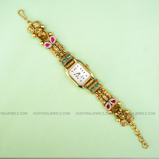 An ornate gold-plated watch bracelet with a rectangular white dial, decorated with pink gemstone accents and small green stone details, shown on a light green background.