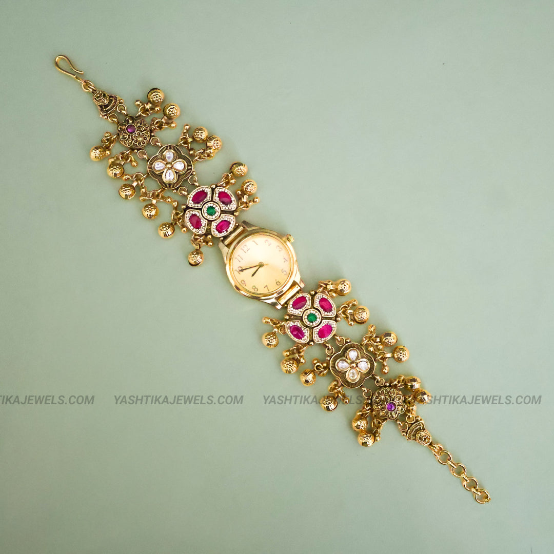 An ornate gold-plated bracelet watch with floral motifs set with pink, green, and white stones, displayed diagonally on a light sage green background.