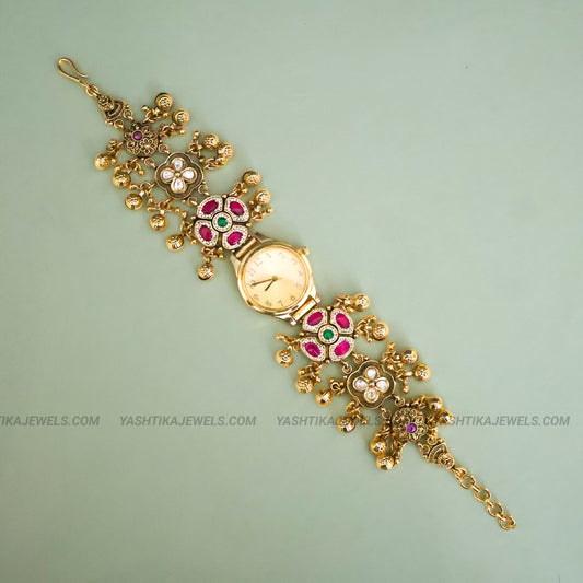 An ornate gold-plated bracelet watch with floral motifs set with pink, green, and white stones, displayed diagonally on a light sage green background.