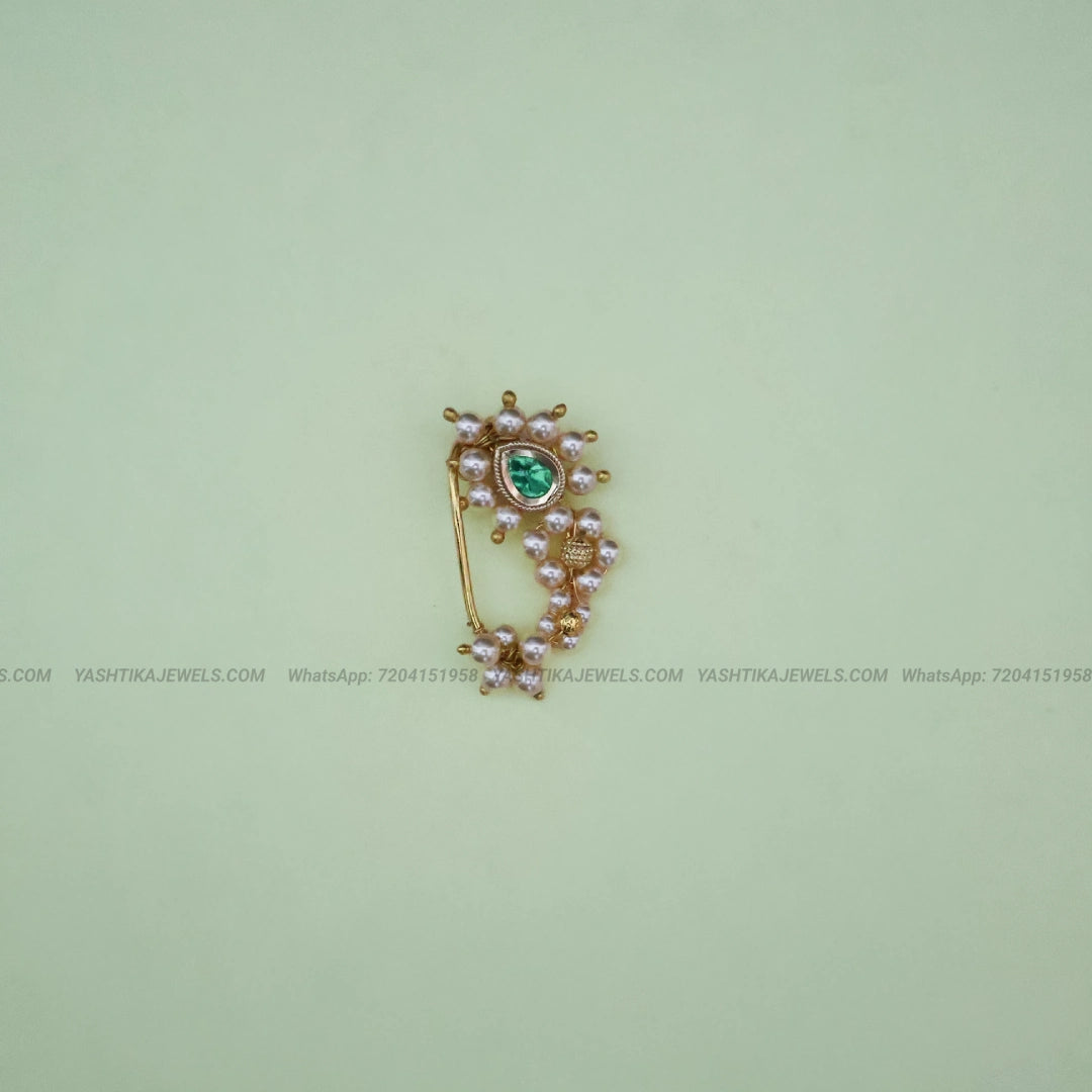 A gold-toned traditional Indian style nose clip featuring a central green stone, surrounded by small pinkish pearl-like beads and gold filigree, set against a pale green background.