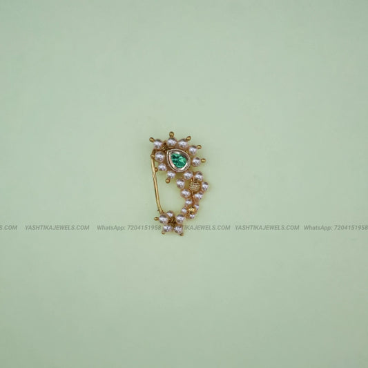 A gold-toned traditional Indian style nose clip featuring a central green stone, surrounded by small pinkish pearl-like beads and gold filigree, set against a pale green background.