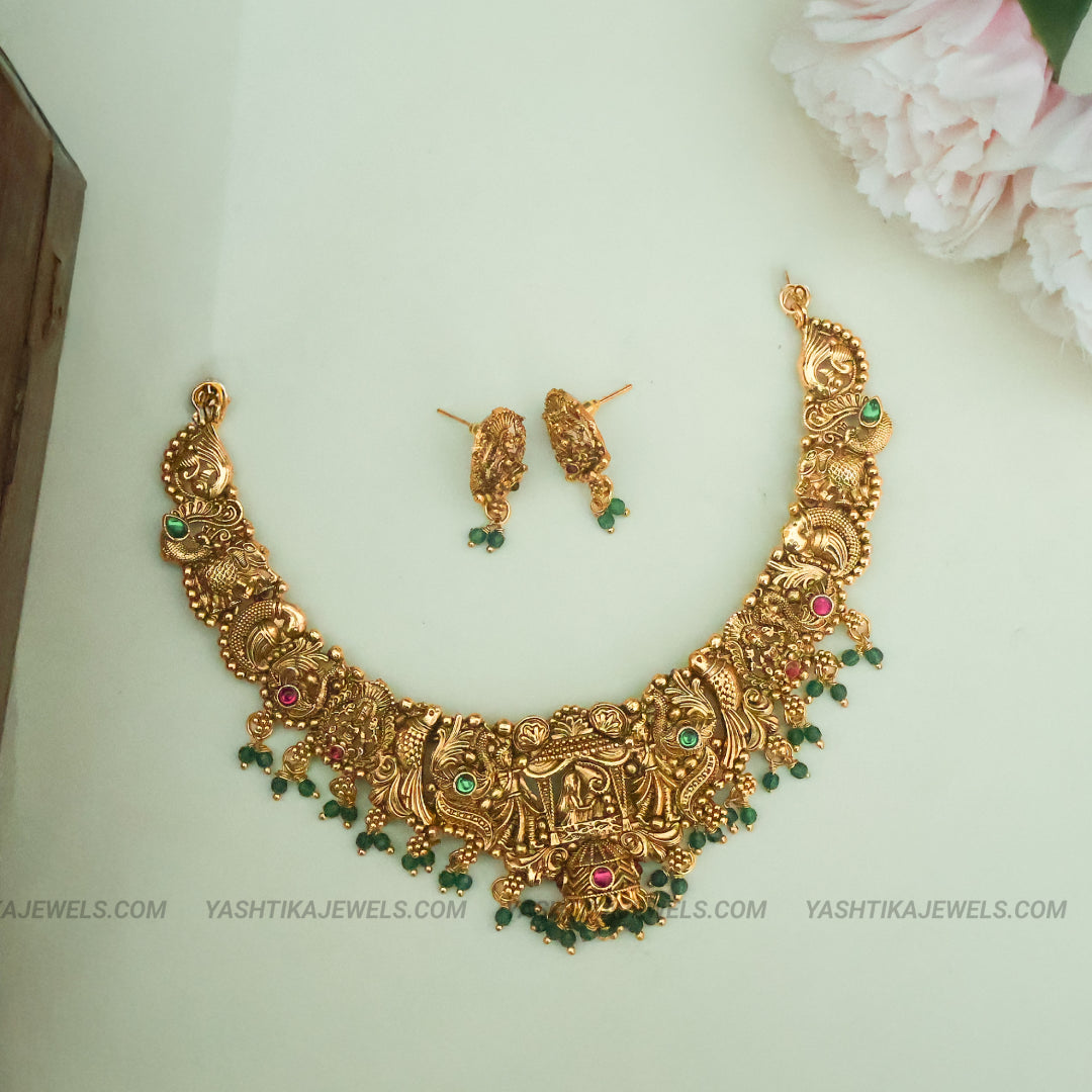 An ornate gold-toned traditional necklace set with matching earrings, decorated with green and pink stones, displayed on a light background with a pink flower in the corner.