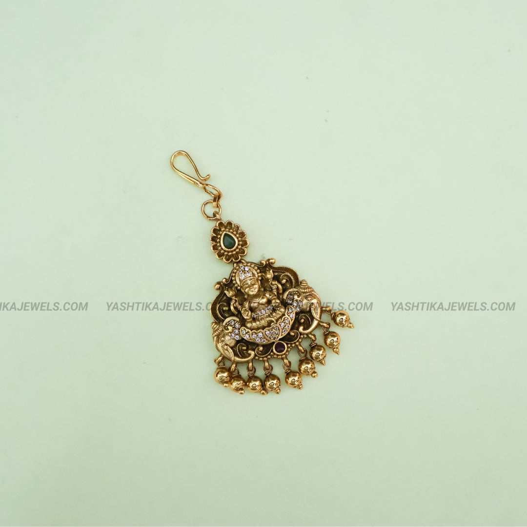 Gold-toned traditional Indian pendant featuring Lord Ganesha, embellished with small green and clear stones, shown on a pale mint green background with watermark text.