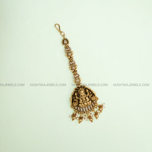 Gold-toned traditional Indian pendant necklace featuring an ornate Ganesha design with small clear stones and dangling pearl-like beads, shown on a pale mint green background.
