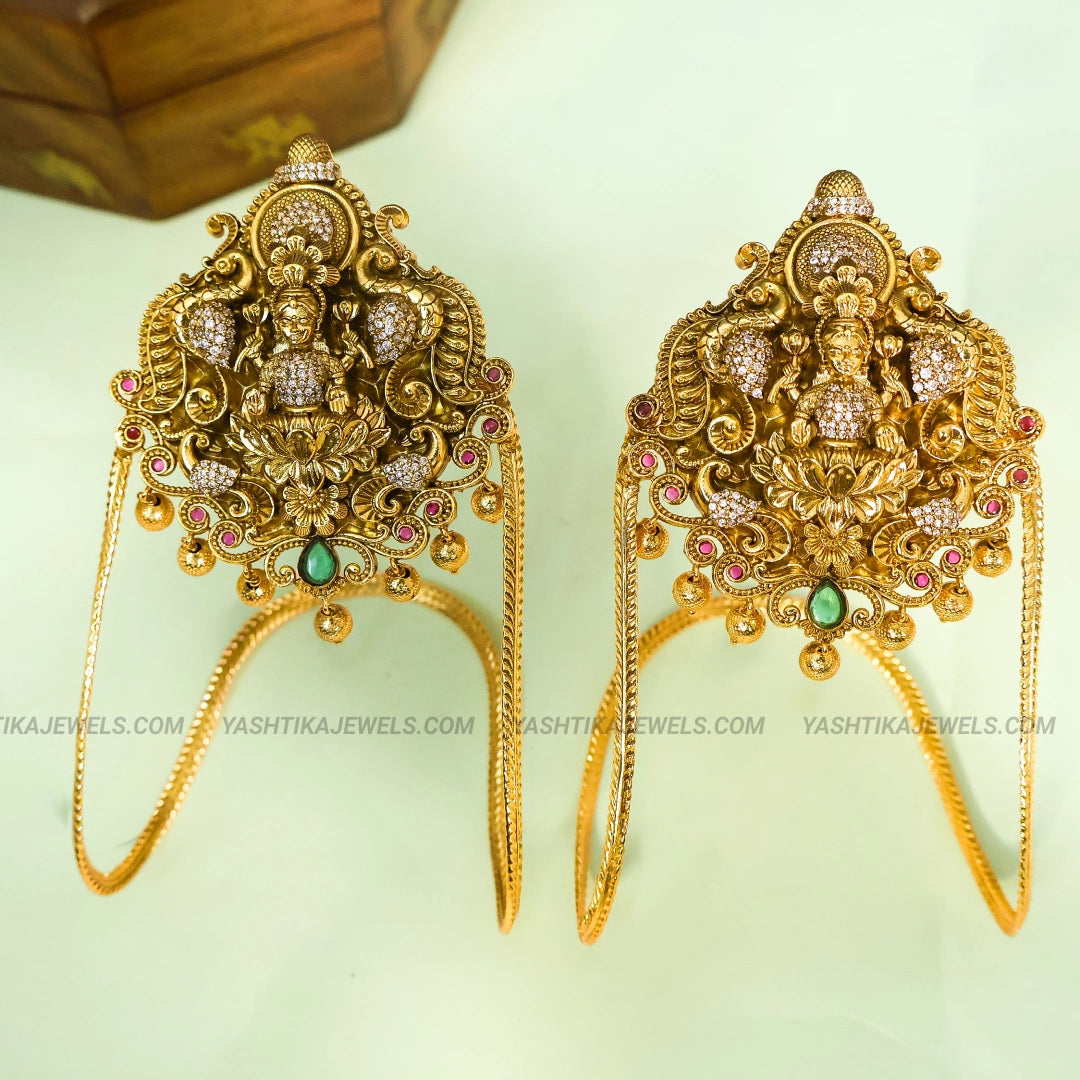 A pair of ornate gold-toned traditional earrings featuring detailed filigree, small crystal-like stones, and green and pink accents, displayed on a pale background with a wooden box partially visible in the upper left.