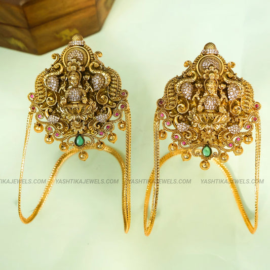 A pair of ornate gold-toned traditional earrings featuring detailed filigree, small crystal-like stones, and green and pink accents, displayed on a pale background with a wooden box partially visible in the upper left.