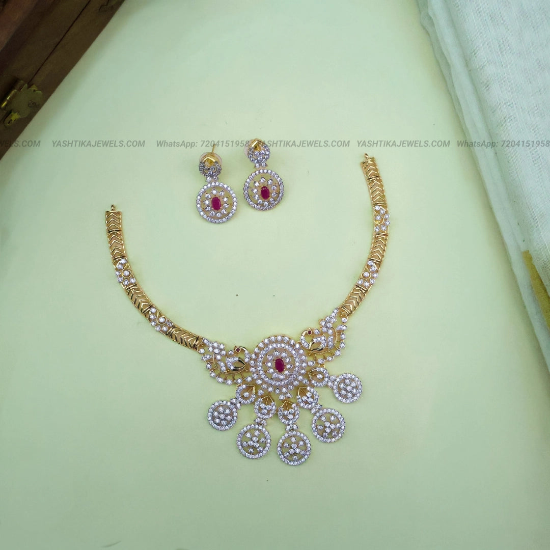 Gold-plated Indian jewelry set featuring a necklace and matching earrings with white stones and red center stones, displayed on a pale green surface.