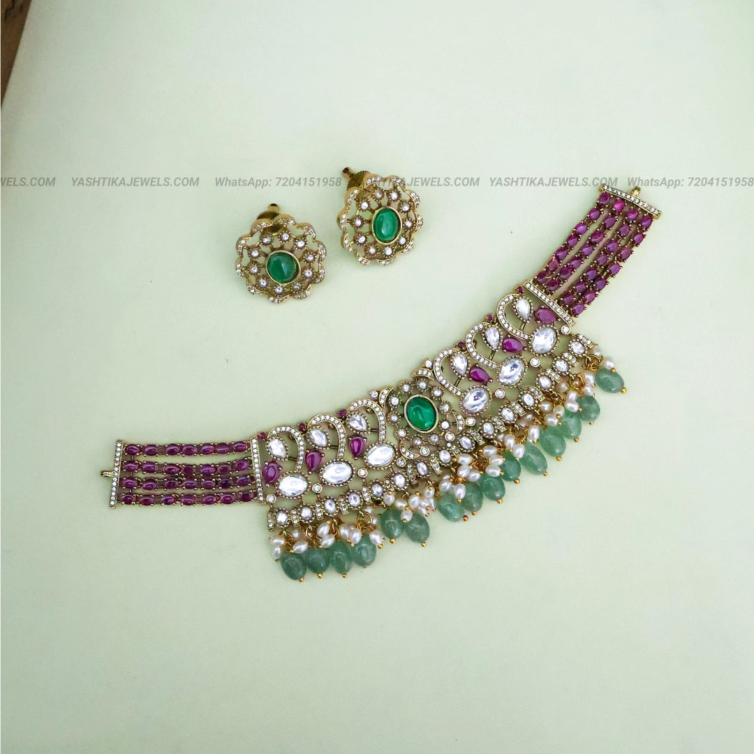 Gold-tone choker necklace set with American diamond stones, ruby-pink stones, green bead drops, pearl detailing and matching earrings.