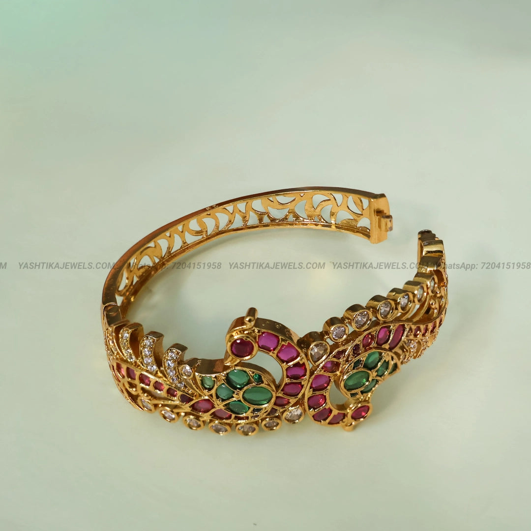 Traditional gold-tone Jadau stone bracelet featuring green and ruby-pink stones, sparkling AD embellishments and intricate peacock-inspired designs.