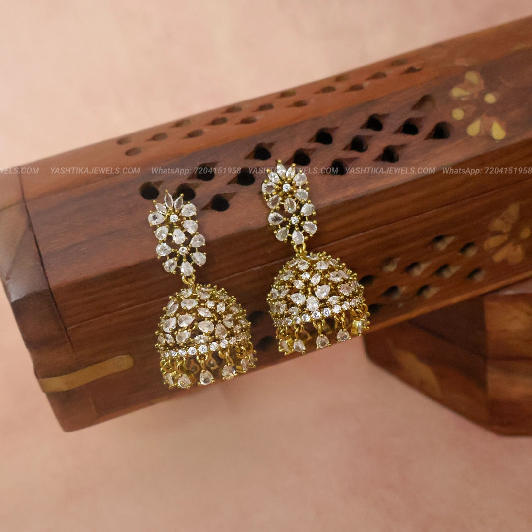 A pair of gold-plated dhamki earrings adorned with clear crystals, displayed on a carved wooden stand against a soft pink background.