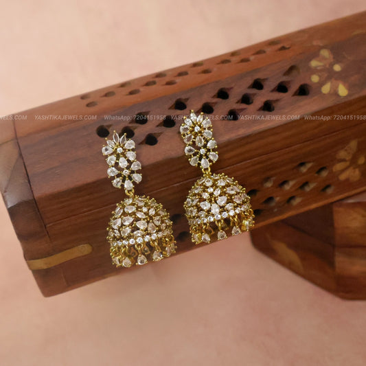 A pair of gold-plated dhamki earrings adorned with clear crystals, displayed on a carved wooden stand against a soft pink background.