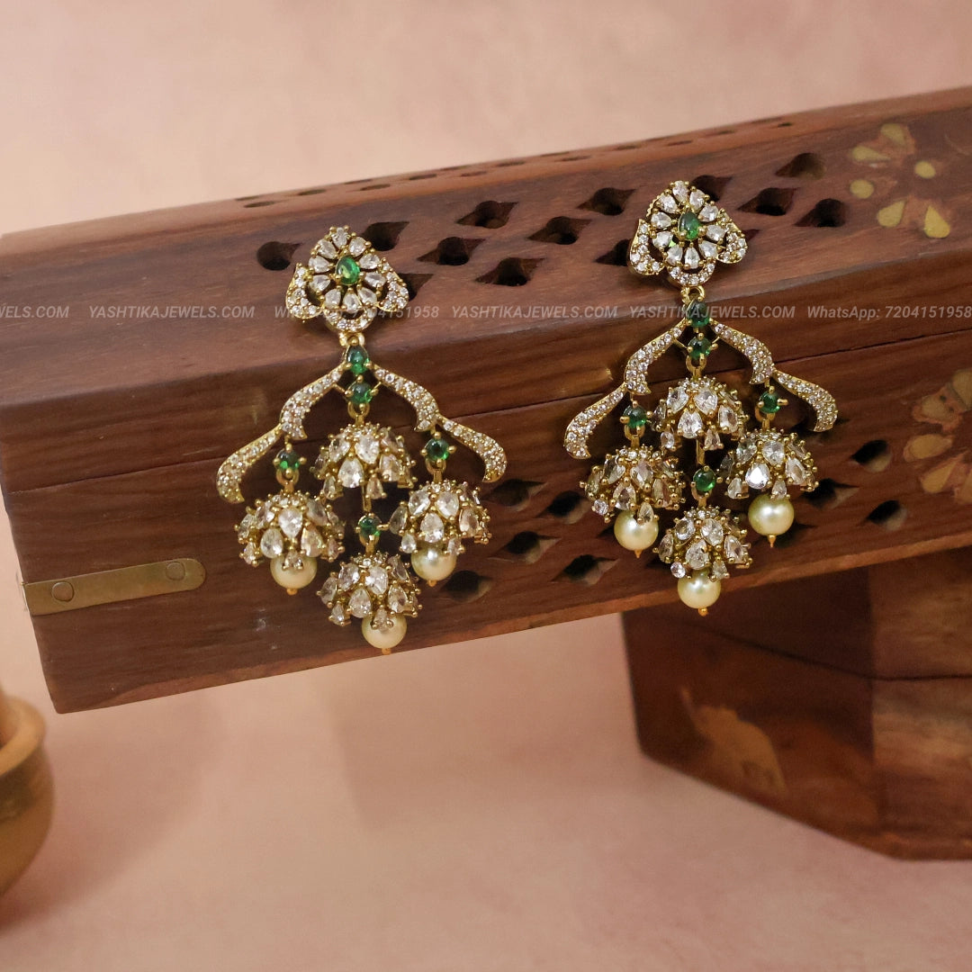 A pair of ornate gold-toned Indian jhumka earrings with green stones and white faux pearls, displayed on a carved wooden stand against a soft pink background.