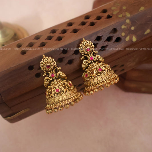 A pair of traditional Indian gold-toned jhumka earrings with small pink gemstone accents, displayed on a carved wooden block against a soft beige background.
