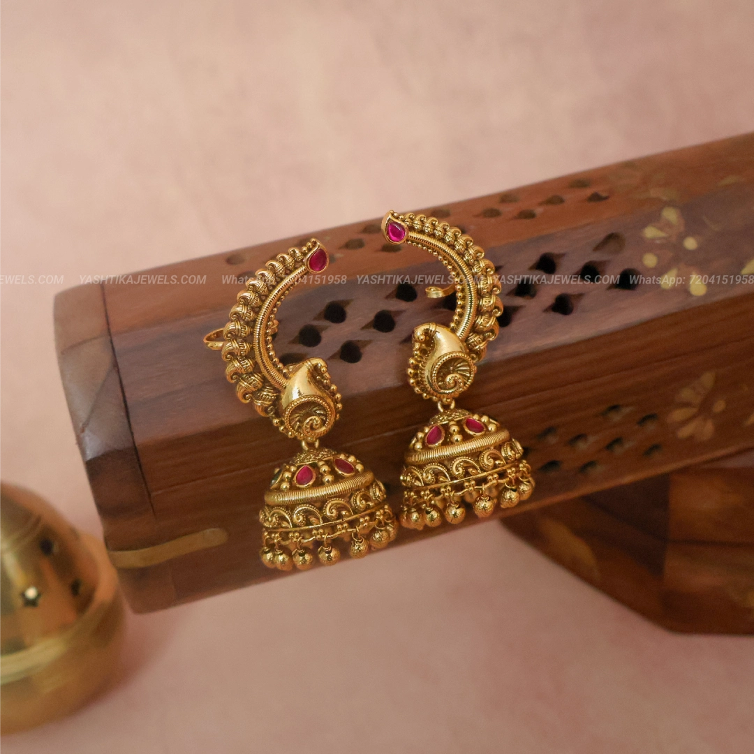 A pair of ornate gold-toned traditional earrings with red gemstone accents, displayed on a carved wooden stand against a soft pink background.