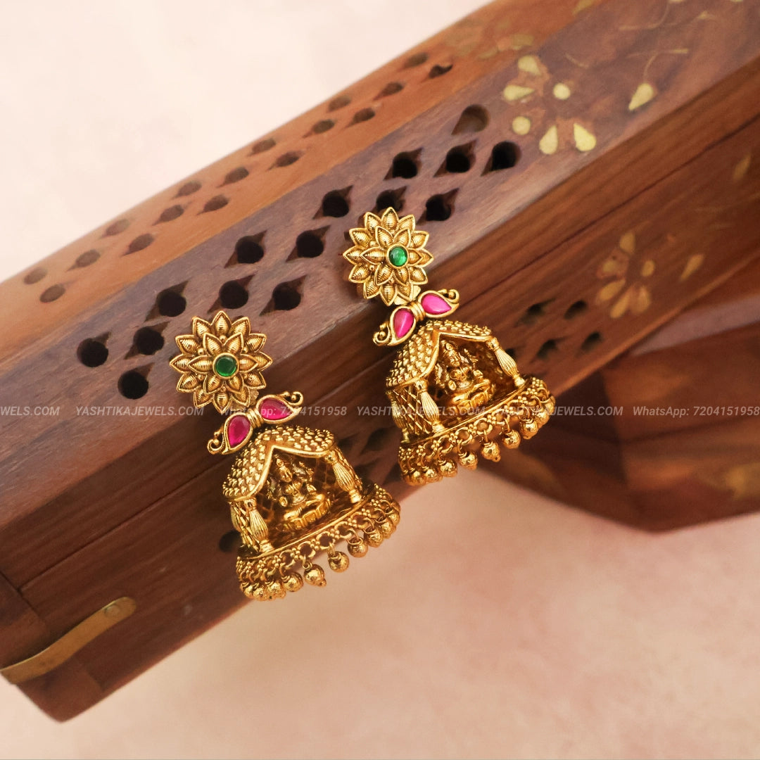 A pair of gold-toned Indian jhumka earrings with green and pink stone accents, displayed on a carved wooden box.