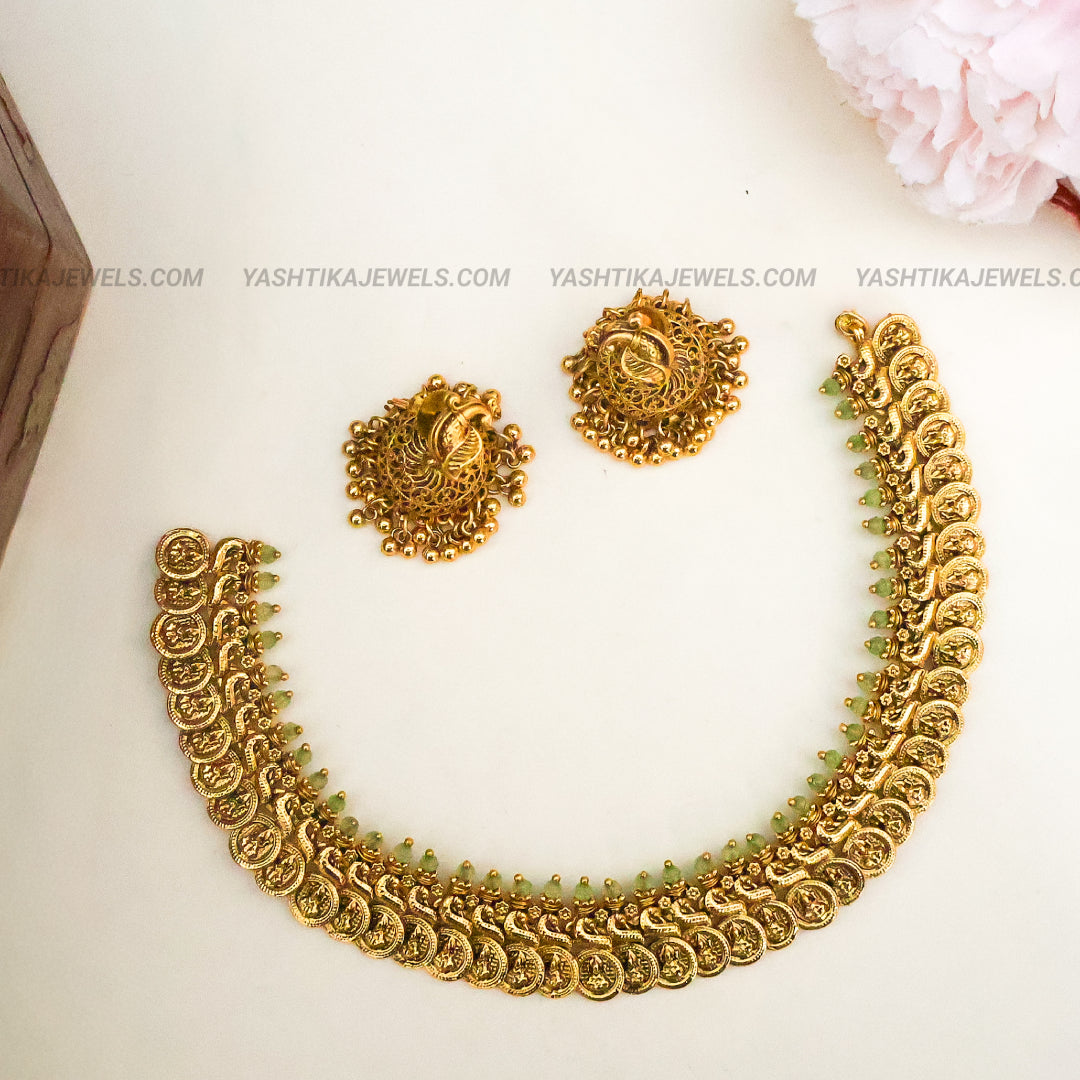 Gold-toned traditional jewelry set featuring a wide scalloped necklace and matching circular earrings with ornate paisley motifs, arranged on a light cream background with a soft pink flower in the corner.